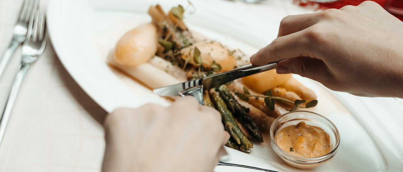 Wellness hotel with gourmet cuisine: Gartenhotel Moser Person cutting asparagus on a plate with sauce at a set dining table