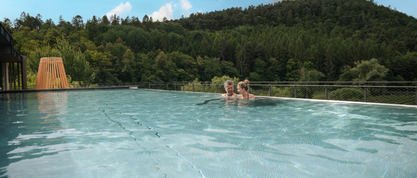 Urlaub in Südtirol im Hotel mit Pool am See Paar entspannt sich im Infinity-Pool mit Aussicht auf bewaldeten Hügel