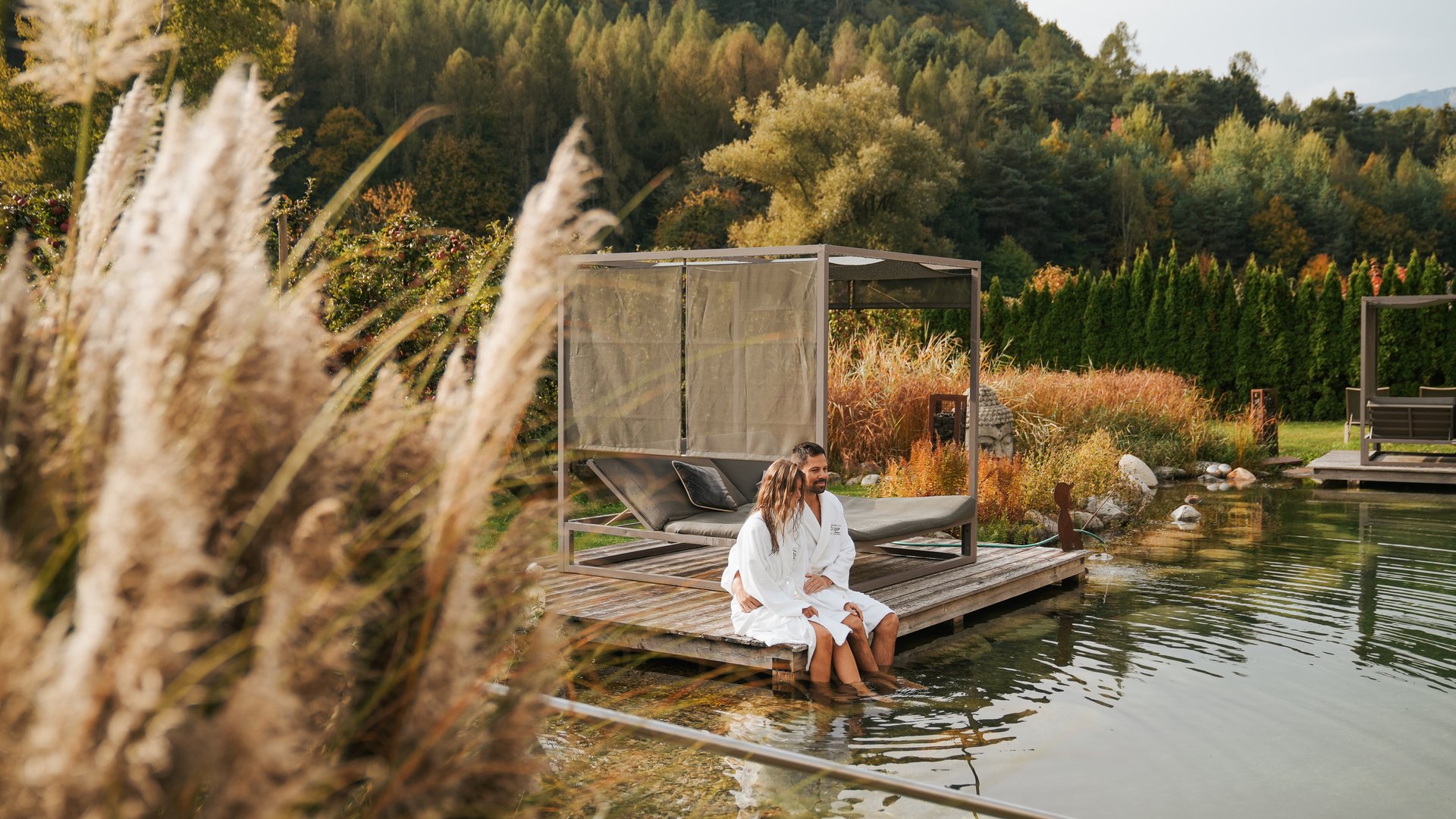 Day spa in South Tyrol: Gartenhotel Moser Couple sitting on wooden pier by natural pond in autumn with mountains behind