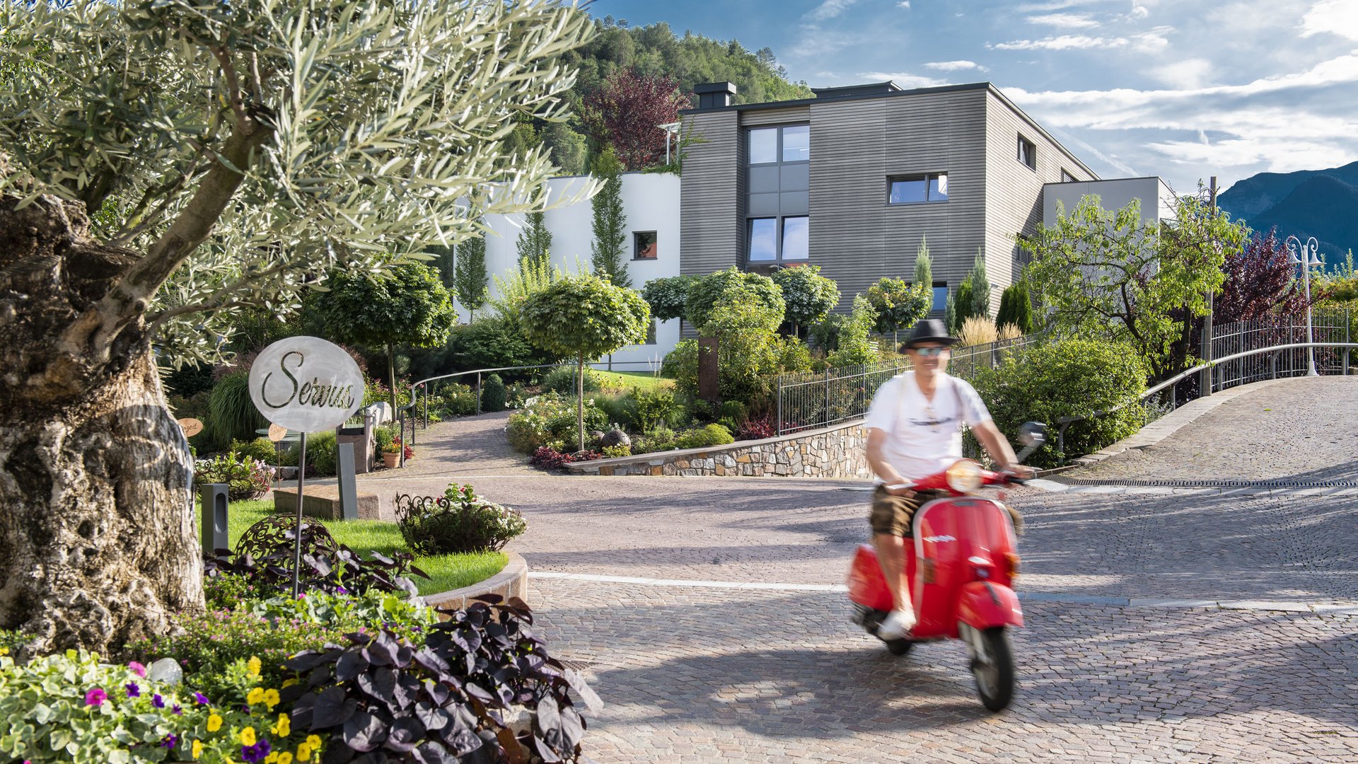 Sporthotel in Südtirol: Gartenhotel Moser Mann fährt roten Roller durch bepflanzte Einfahrt vor modernem Haus