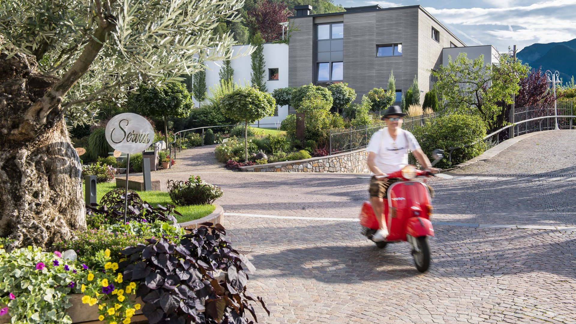 Hotel by the lake in South Tyrol: cabriolet & Vespa Man riding red scooter through landscaped driveway in front of modern house