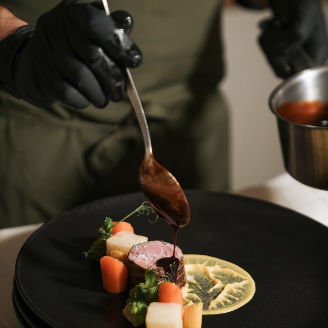 Im Wellnesshotel glutenfrei genießen Koch gießt Sauce auf Fleischgericht mit Gemüse auf schwarzem Teller