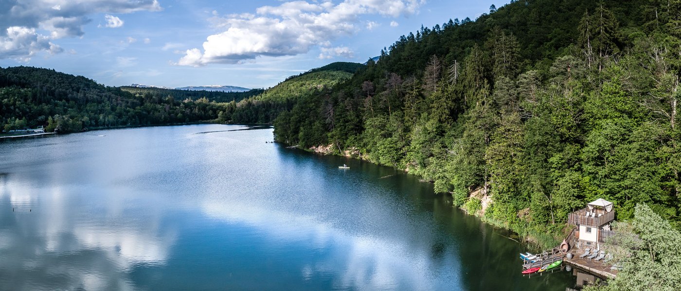 Lago di Caldaro: passeggiata e varie attività nei dintorni del Gartenhotel Moser Lago con riva boscosa e piccolo molo sotto un cielo blu con nuvole