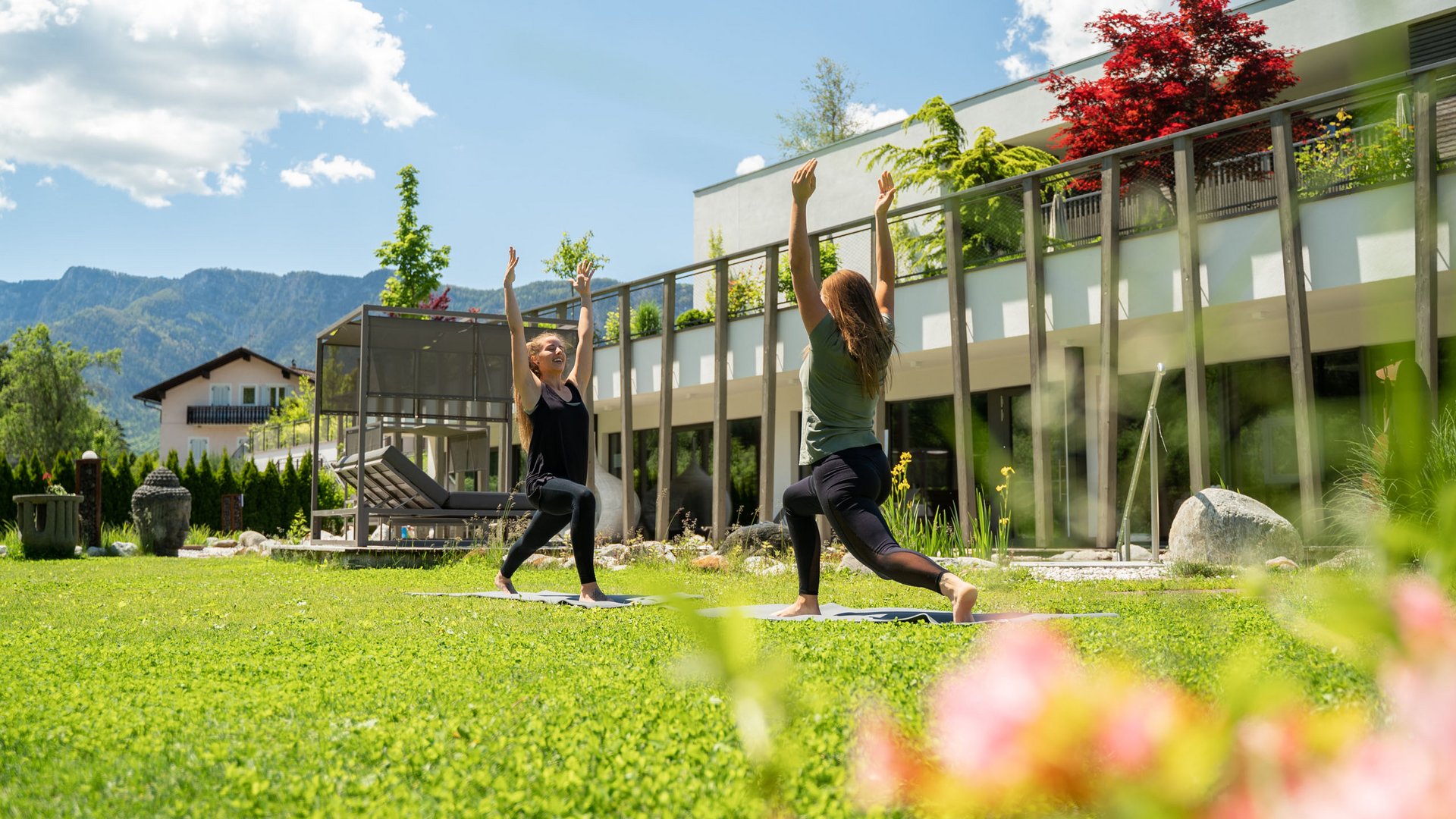 Yoga in Alto Adige Due donne fanno yoga sull'erba davanti a un edificio moderno sotto il sole