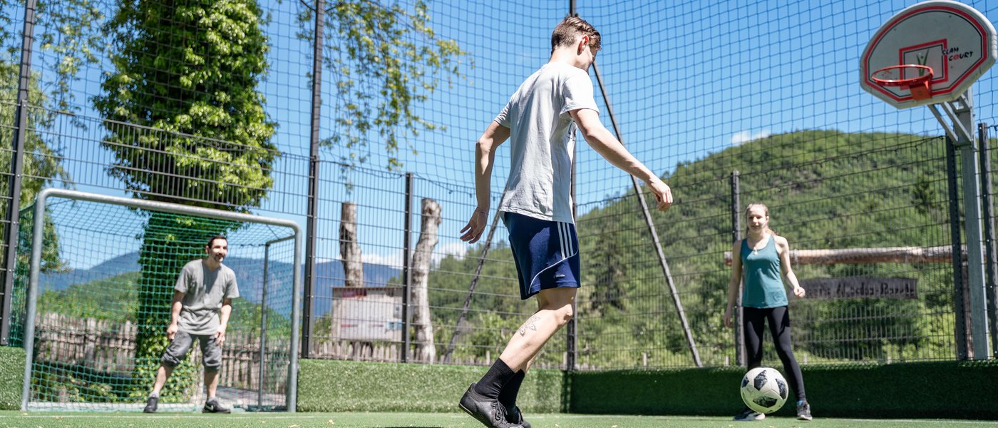Familienhotel in Südtirol: Gartenhotel Moser Drei Personen spielen Fußball auf einem grünen eingezäunten Sportplatz bei sonnigem Wetter