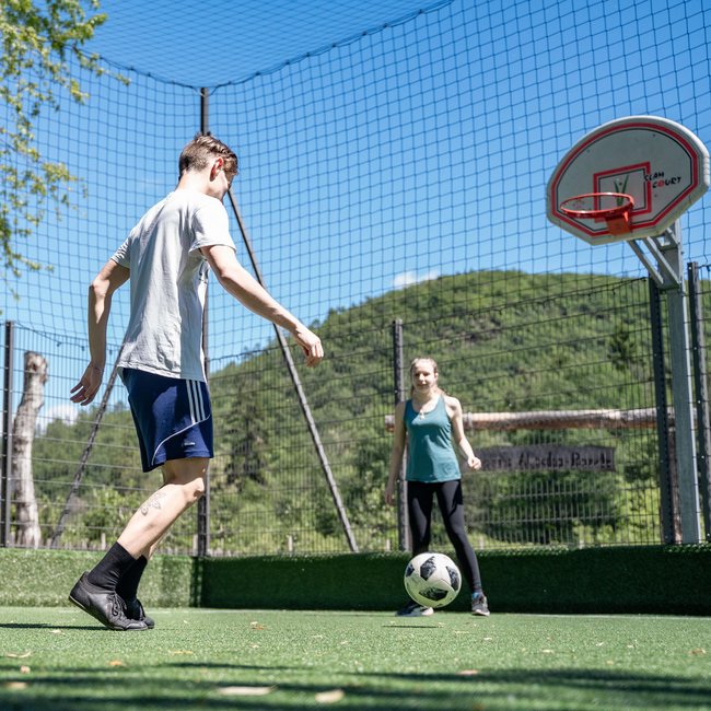 Sporthotel in Südtirol: Gartenhotel Moser Drei Personen spielen Fußball auf einem grünen eingezäunten Sportplatz bei sonnigem Wetter