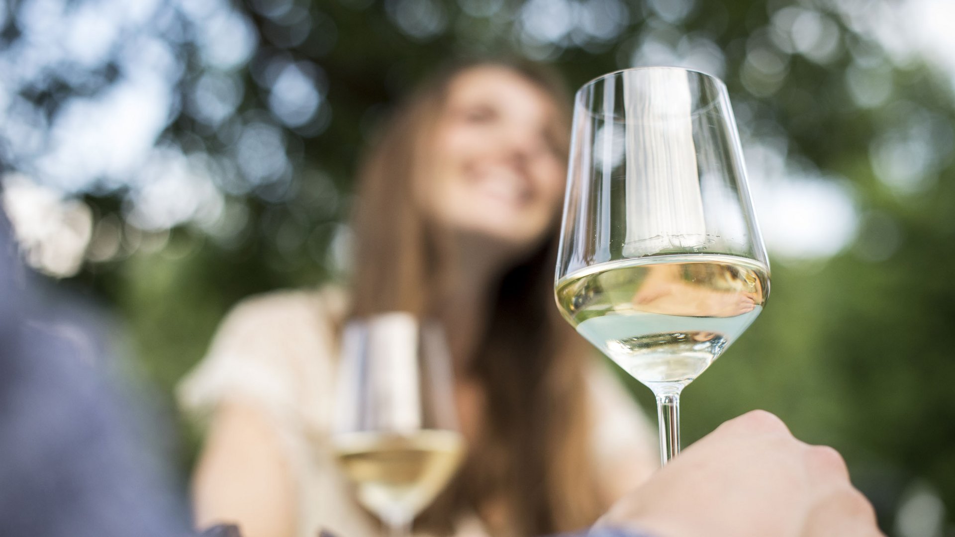 Hotel sulla Strada del Vino in Alto Adige: Gartenhotel Moser Bicchiere di vino bianco in primo piano, donna sorridente sfocata sullo sfondo