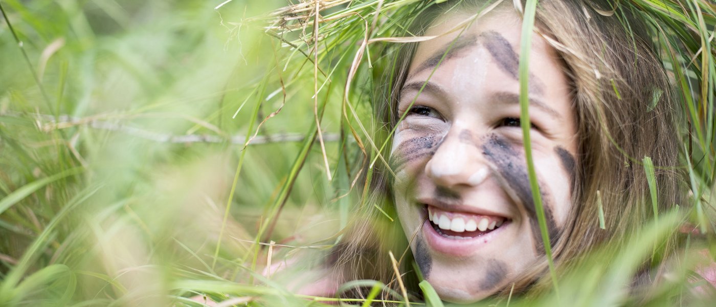 Familienhotel in Südtirol: Gartenhotel Moser Lachendes Mädchen mit Tarnfarbe und Graskranz im Gesicht im Freien