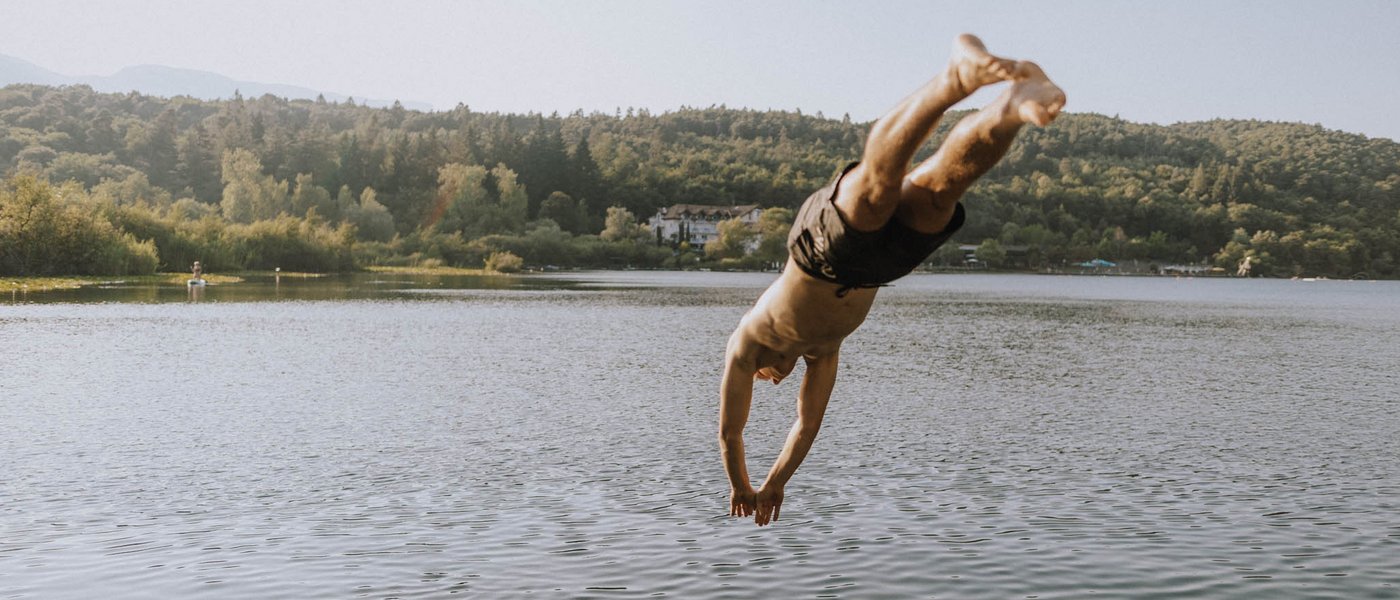 Hotels nahe dem Kalterer See 4-Sterne-Superior Mann taucht von Steg in ruhigen See vor bewaldetem Hügel