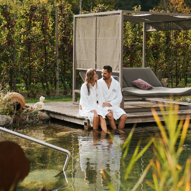 Hotel am Montiggler See: Gartenhotel Moser Paar in Bademänteln sitzt am Steg eines natürlichen Pools umgeben von Natur