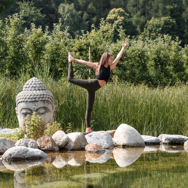 Hotels bei Eppan: Gartenhotel Moser Frau macht Yoga-Pose neben Buddha-Skulptur an einem See in der Natur