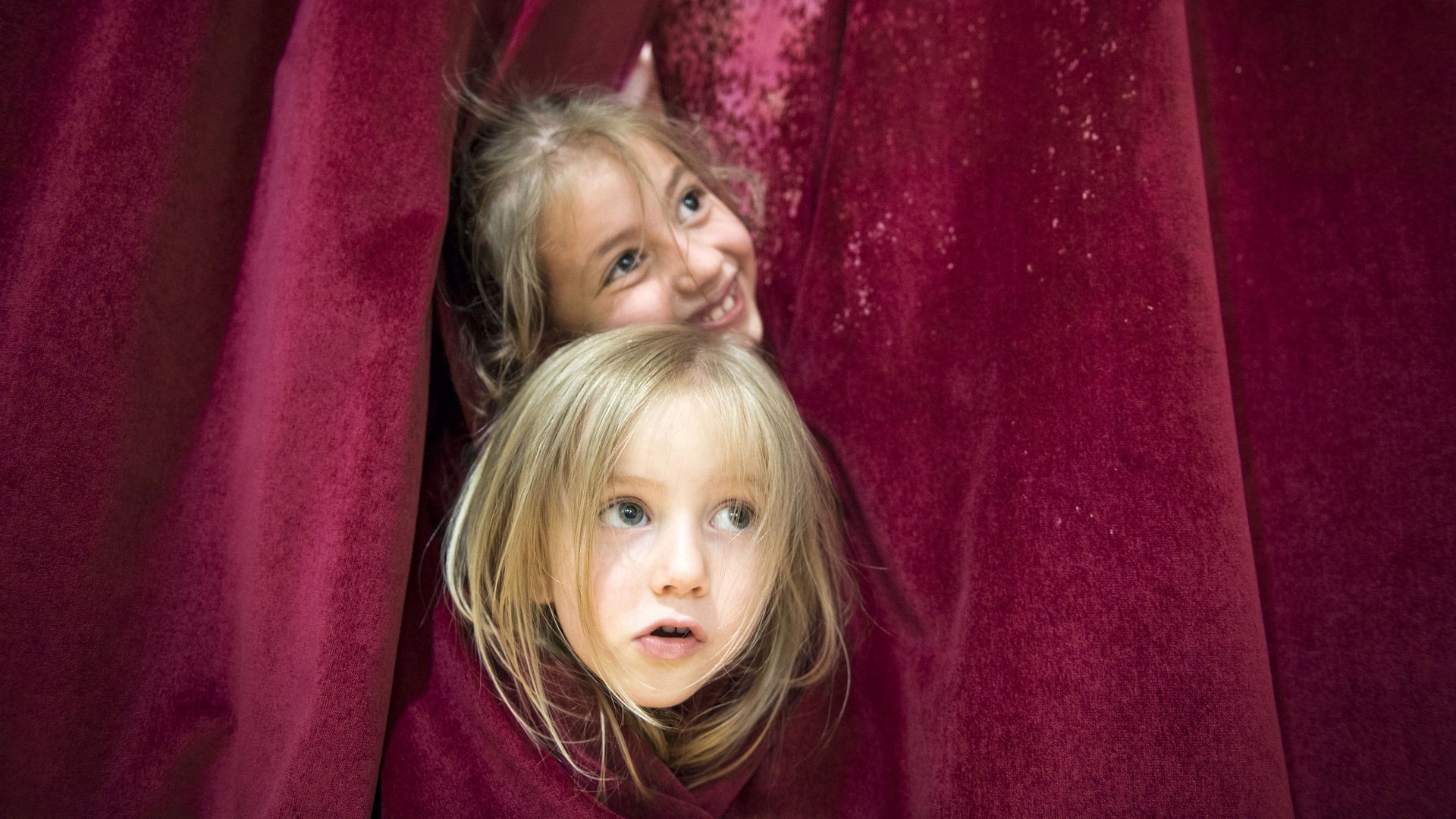 Children’s hotels in South Tyrol with 4-star-S service Two children peeking curiously from red curtains