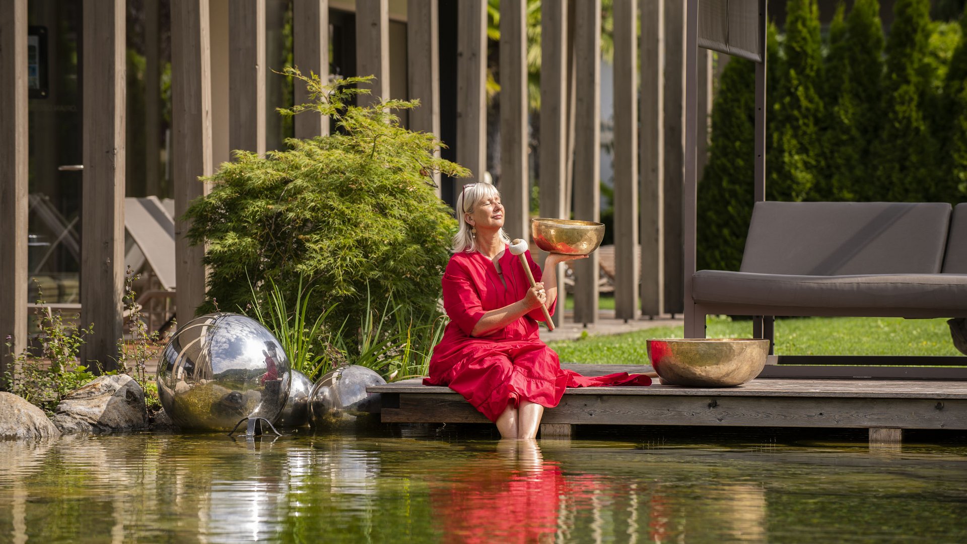 Hotel am Montiggler See: Gartenhotel Moser Frau in rotem Kleid sitzt am Teichrand mit Klangschale und schlägt sie an