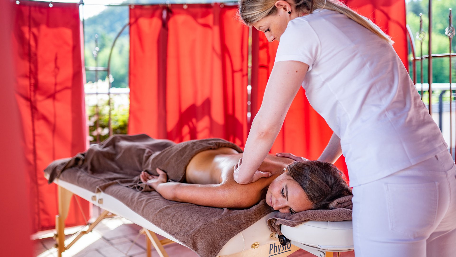 Hotel am Montiggler See: Gartenhotel Moser Frau erhält Massage auf Liege in entspannter Umgebung mit roten Vorhängen