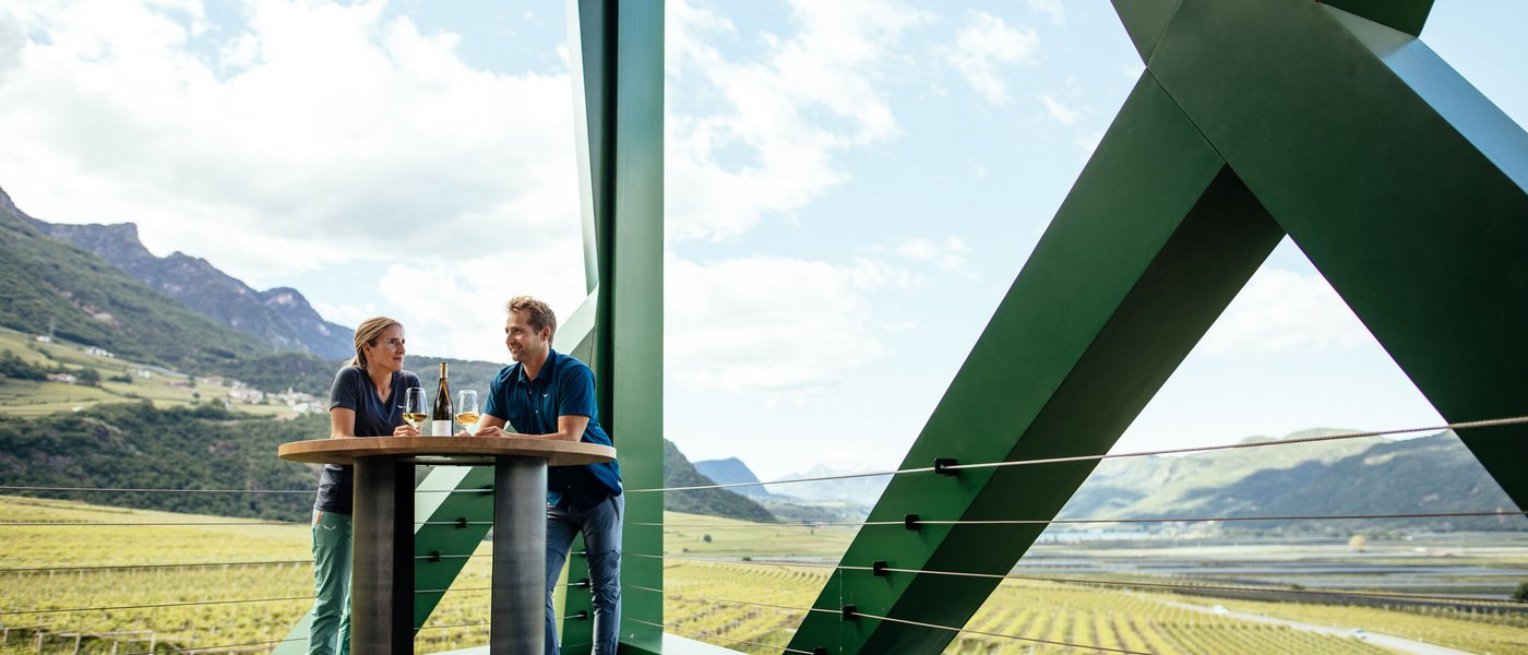 Hotels on Lago di Monticolo: South Tyrol awaits Two people at a standing table with wine overlooking mountains and vineyards