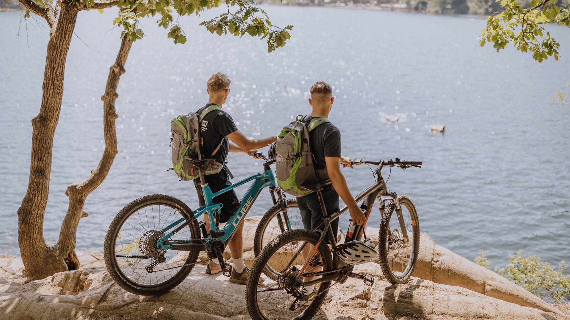 Wellnessurlaub am See: Gartenhotel Moser Zwei Radfahrer mit Rucksäcken stehen an einem Seeufer unter Bäumen