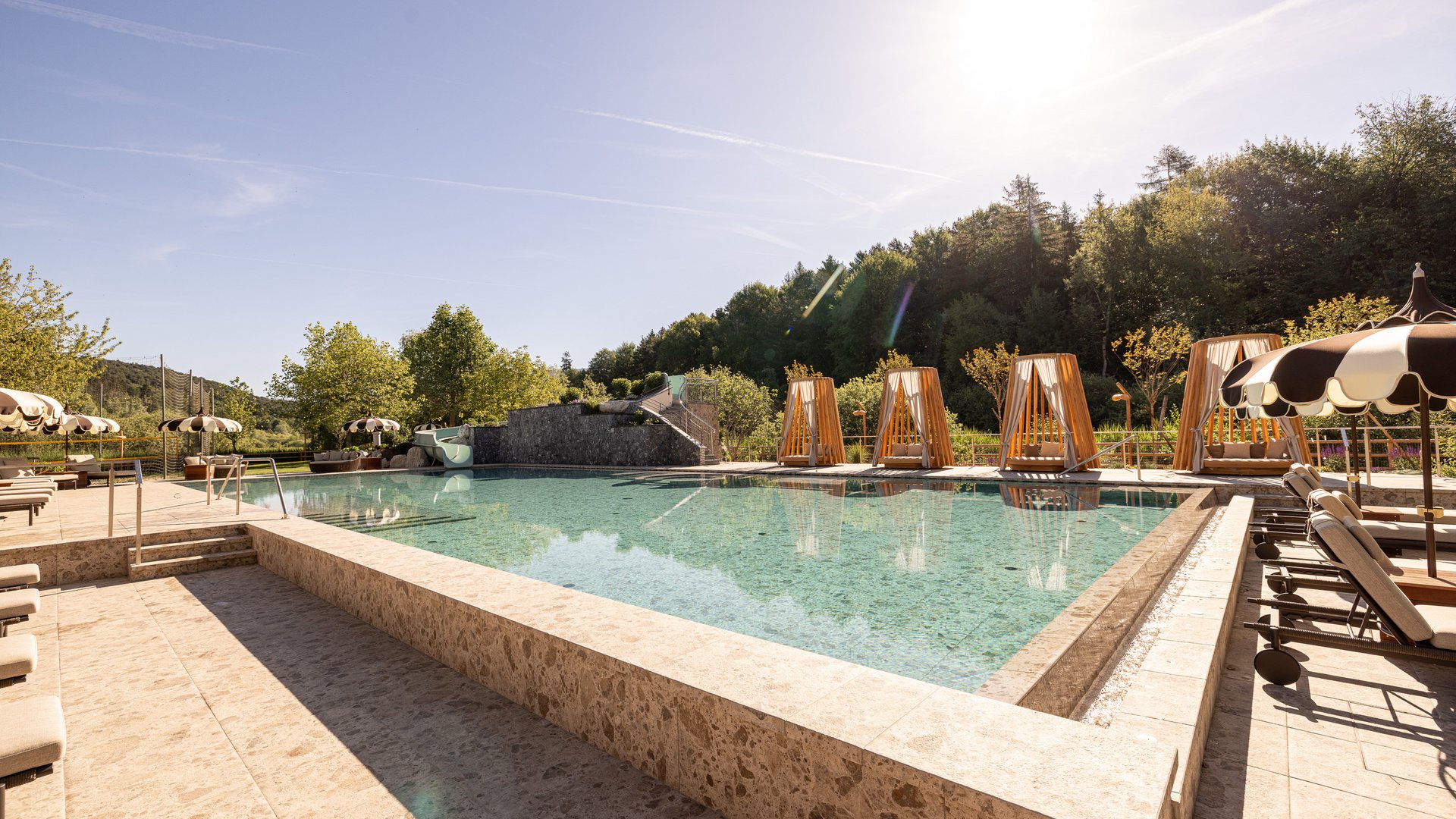 Novità a partire dal 2026 al Gartenhotel Moser Piscina all'aperto con lettini, ombrelloni e cabine in legno sotto il sole
