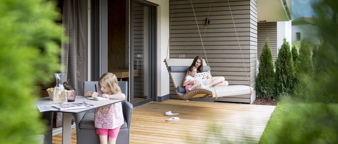 Familienhotel in Südtirol: Gartenhotel Moser Kind malt am Tisch auf Terrasse, Mutter liest Kind auf Hängeliege vor