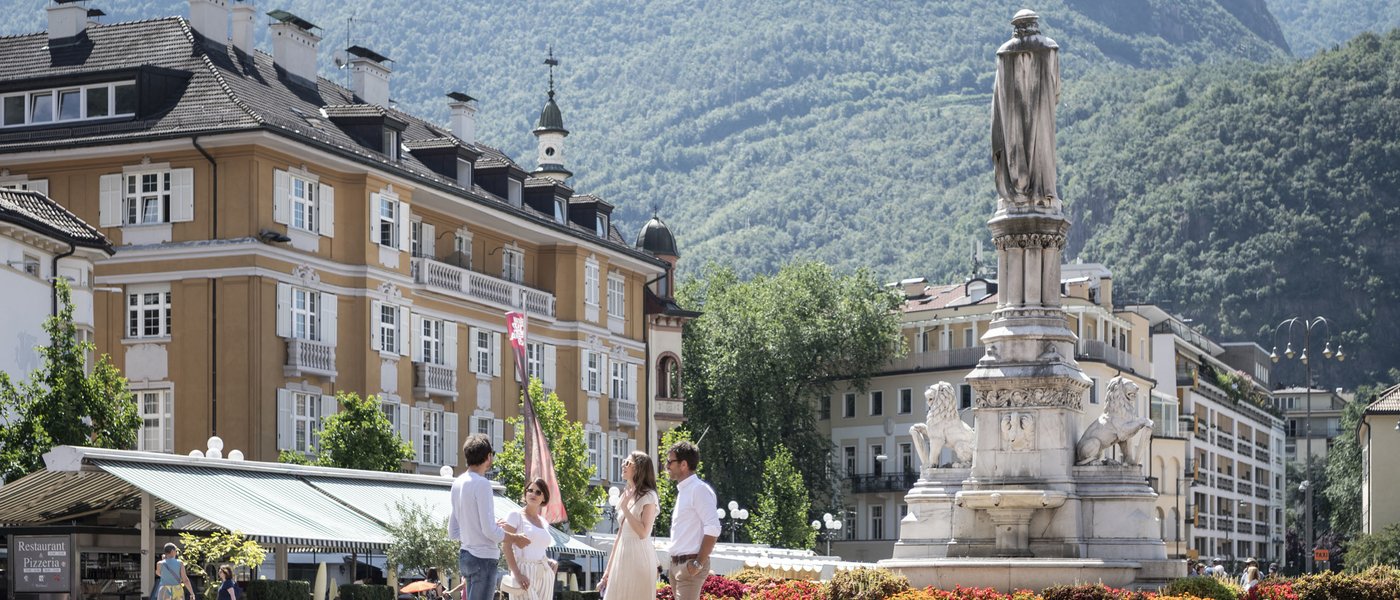 Hotels on Lago di Monticolo: South Tyrol awaits People standing in historic square with statue and mountains in the background