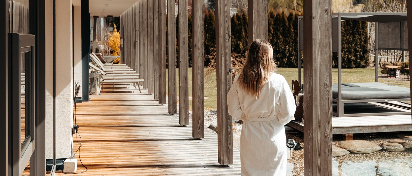 Urlaub in Südtirol im Hotel mit Pool am See Frau in Bademantel auf Holzveranda neben Pool und Garten im Sonnenschein