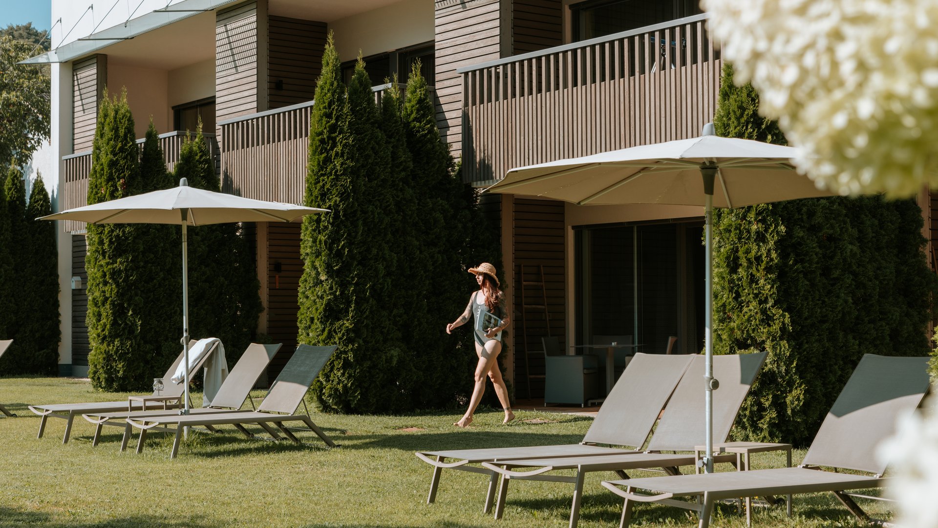 Hotels nahe dem Kalterer See 4-Sterne-Superior Frau mit Sonnenhut geht neben Liegestühlen und Sonnenschirmen im Garten von Hotelanlage