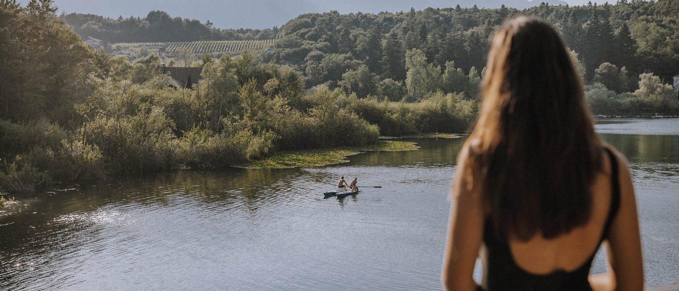 Laghi balneabili in Alto Adige: cosa c’è da sapere Donna guarda fiume con due persone in barca a remi davanti a colline boschive