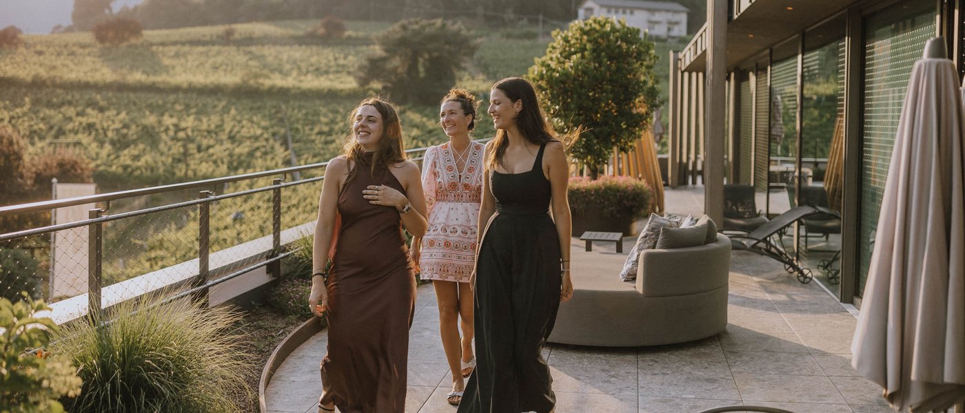 Laghi balneabili in Alto Adige: cosa c’è da sapere Tre donne che passeggiano su una terrazza con vista su montagne e boschi al tramonto