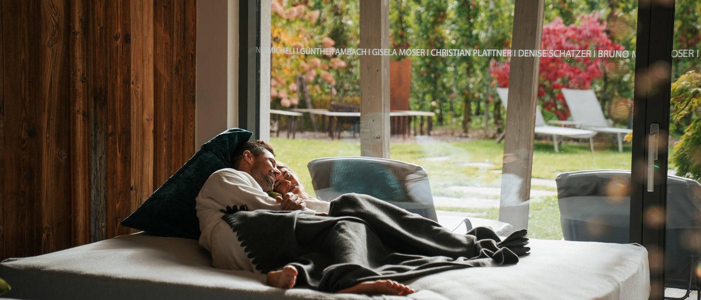 Wellnesshotel mit Whirlpool und Saunawelt Paar liegt gemütlich auf einem Bett vor großer Glasfront mit Gartenblick