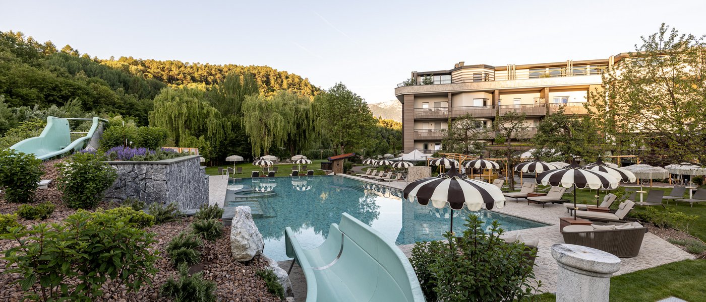 Familienhotel in Südtirol: Gartenhotel Moser Außenpool mit Wasserrutsche, Liegestühlen und Sonnenschirmen vor Hotel und Wald