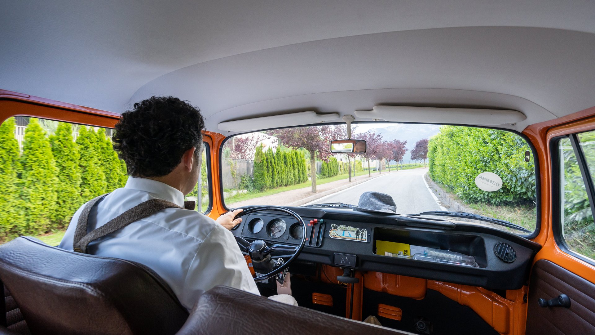 Hotels in Eppan, Südtirol: Anfahrt Mann fährt einen orangefarbenen Oldtimer auf einer Straße mit grünem Buschwerk