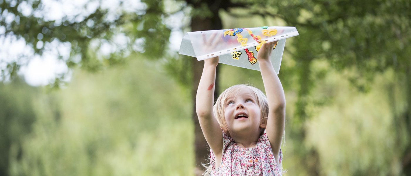 Familienhotel in Südtirol: Gartenhotel Moser Kind hebt ein buntes Papierflugzeug hoch im Freien unter Bäumen